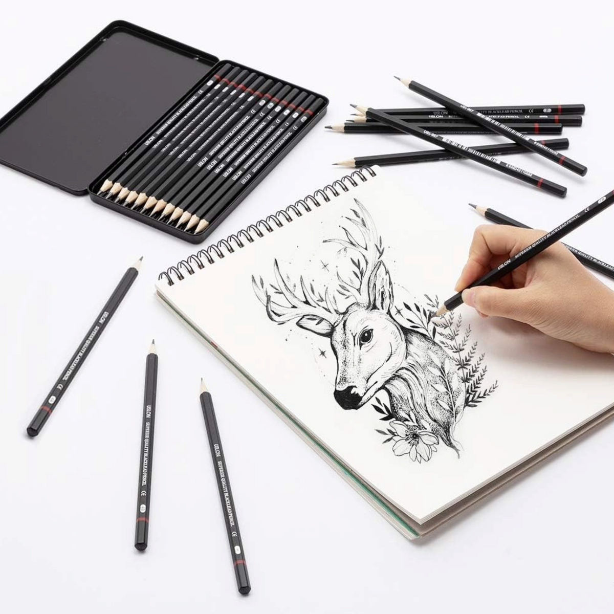 Elite Stationery House 12-piece graphite sketching pencil set in a metal tin with a hand drawing a deer illustration.